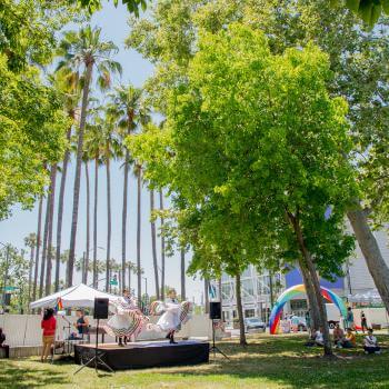 A pair of dancers wearing white traditional Mexican dresses with colorful ruffles perform on a small black stage in a park with tall green trees and festival booths in the background