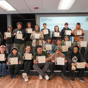 21 young adults sit and stand for a group photo in a classroom setting, holding up certificates and smiling at the camera 