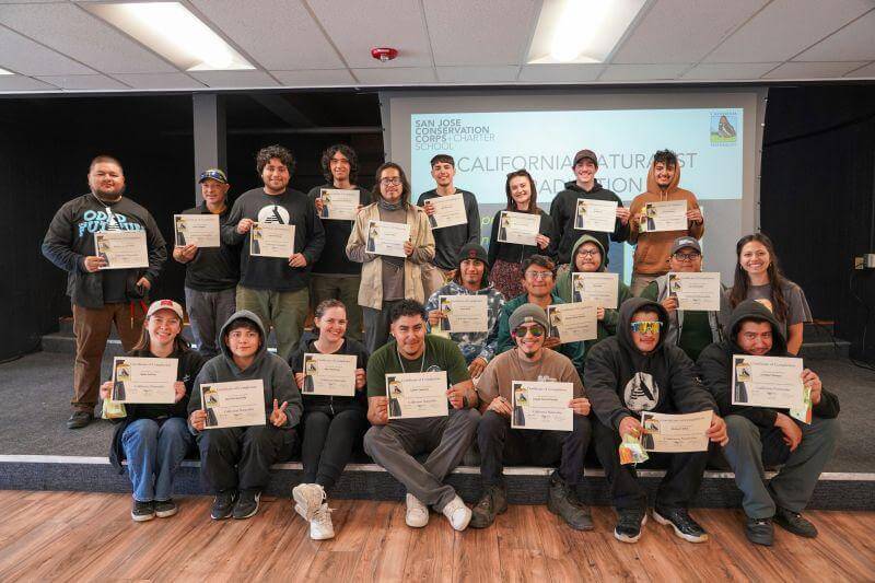 21 young adults sit and stand for a group photo in a classroom setting, holding up certificates and smiling at the camera 
