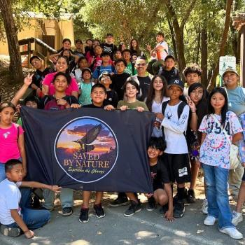 A group of about 30 middle school aged students stand in a natural park area facing the camera and holding up a large dark flag with a circular emblem with the Saved By Nature logo of an eagle and sunset