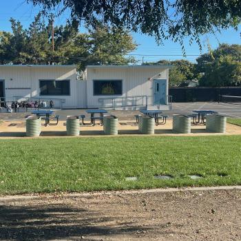 A row of six raised metal garden beds next to four school picnic tables surrounded by green grass, in front of a school blacktop with portable classrooms behind them