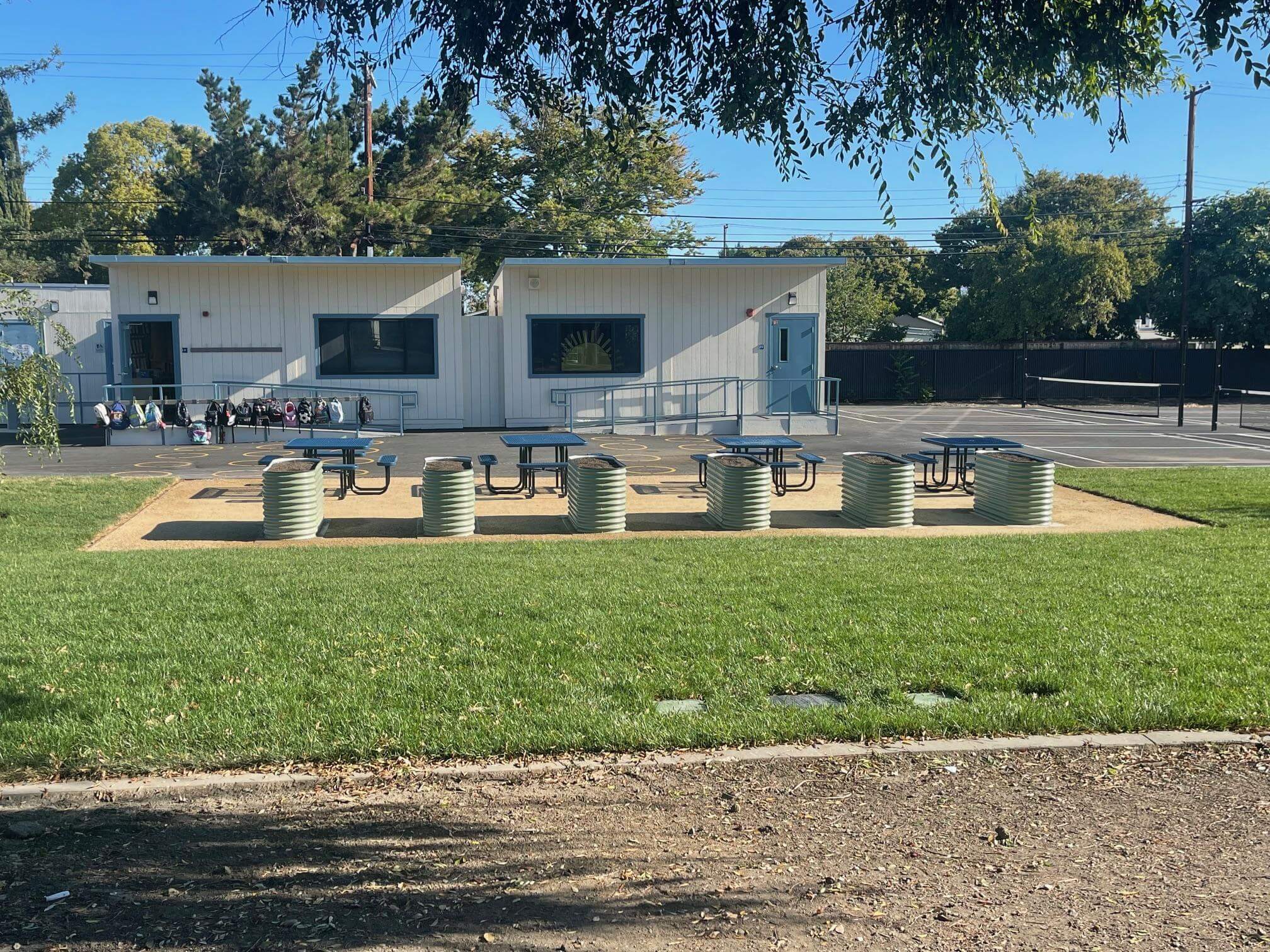 A row of six raised metal garden beds next to four school picnic tables surrounded by green grass, in front of a school blacktop with portable classrooms behind them