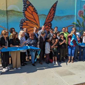 A large group of adults and children stand in front of a large colorful mural with a monarch butterfly, behind a recently cut blue ceremonial ribbon