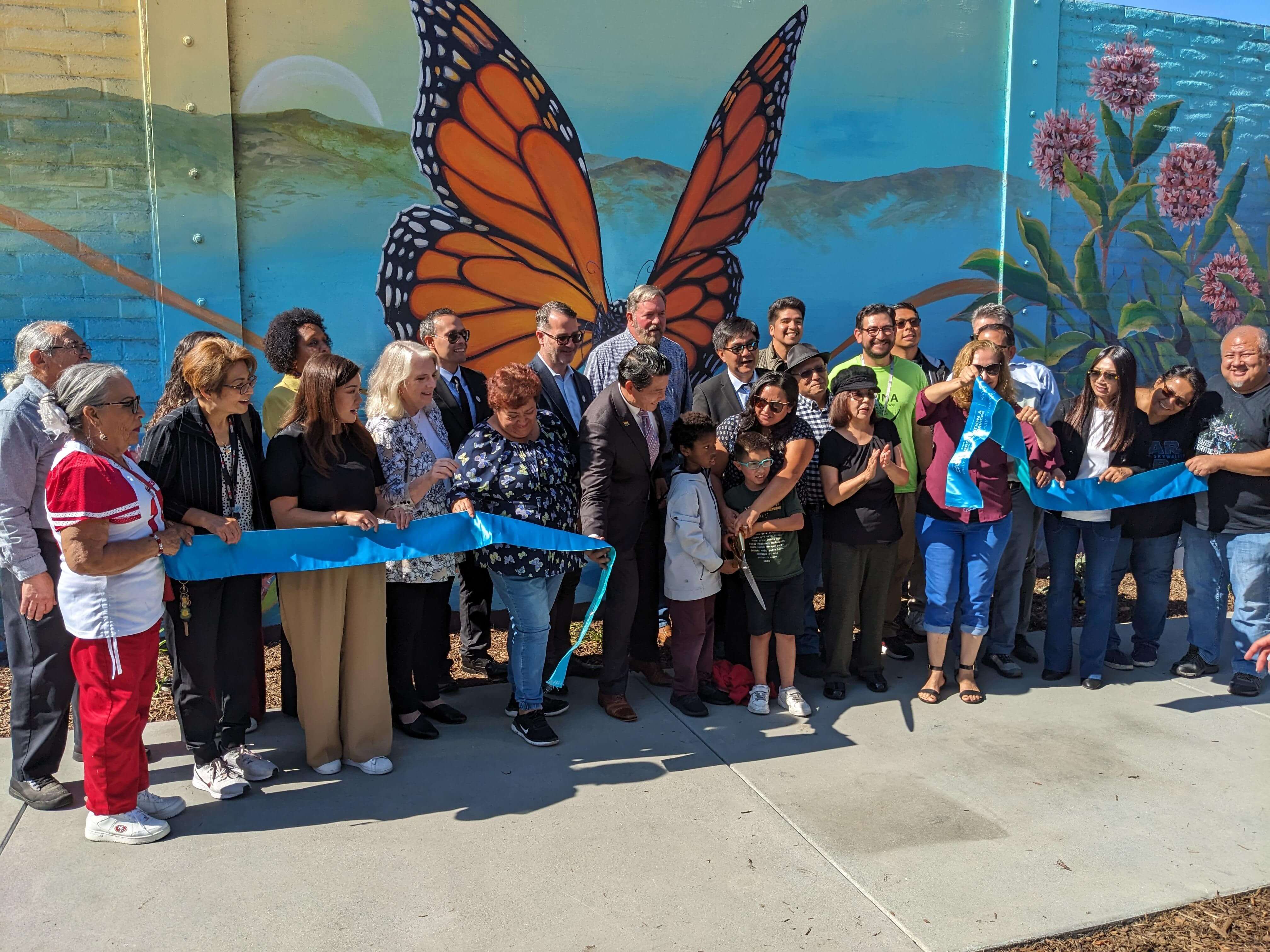 A large group of adults and children stand in front of a large colorful mural with a monarch butterfly, behind a recently cut blue ceremonial ribbon