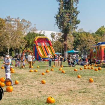 Photograph of a vibrant Halloween event in a park, featuring a colorful inflatable slide, pumpkins scattered on grass, and food trucks with people milling about.