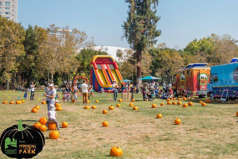 Photograph of a vibrant Halloween event in a park, featuring a colorful inflatable slide, pumpkins scattered on grass, and food trucks with people milling about.