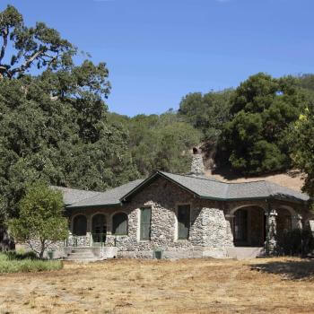 A single-story stone house with a gray shingled roof, surrounded by lush green trees and set on a dry, yellowed grassy field. The house has a small porch with columns and multiple windows, situated in a rural, hilly area under a clear blue sky.