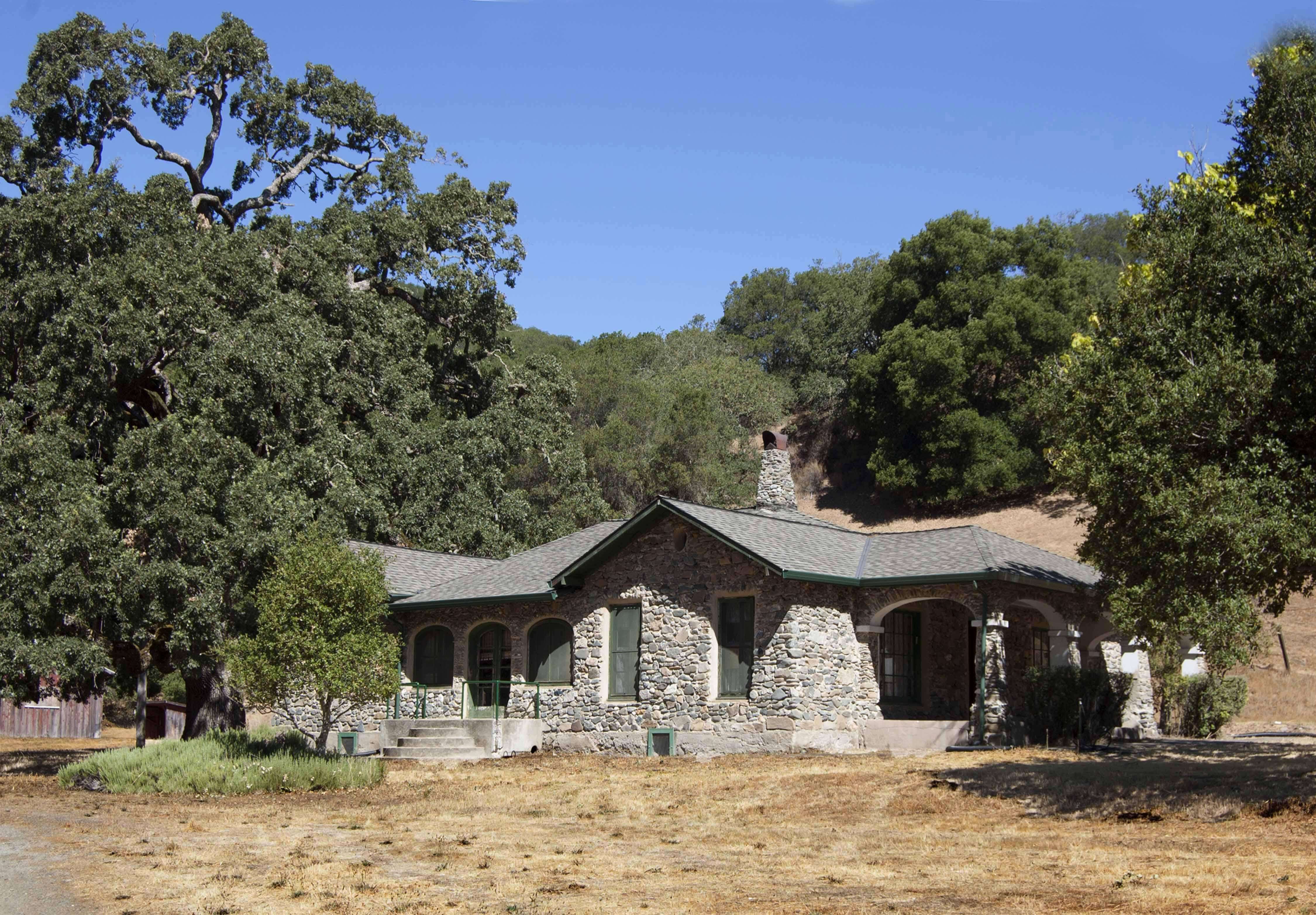 A single-story stone house with a gray shingled roof, surrounded by lush green trees and set on a dry, yellowed grassy field. The house has a small porch with columns and multiple windows, situated in a rural, hilly area under a clear blue sky.
