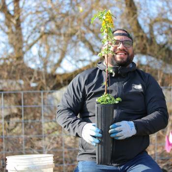 A bearded man with glasses, wearing a black jacket and blue gloves, smiling while holding a black plant pot with green plants and yellow flowers. 
