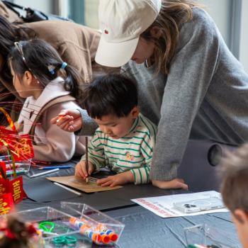 Photograph of three children and adults, one woman in a grey sweater and white cap, helping a boy with a coloring page at a table with colorful activities and toys.