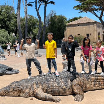 Five children standing on large, life-sized crocodile statue in a sunny park with trees, people, and a stone building in the background.