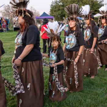 Group of members from Muwekma Ohlone Tribe in traditional dress with feathered headdresses walking in a cultural festival procession outdoors.