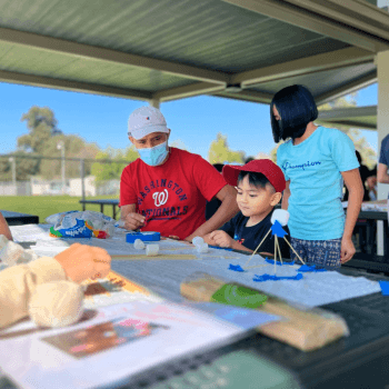 A boy in a red hat sitting at a covered picnic table with two adults working on a craft activity