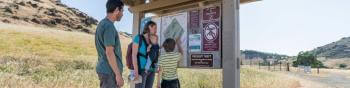 A family of four, two adults, a child, and a baby being carried by the mother, gather around a small trail kiosk with a large map and other informational signage.