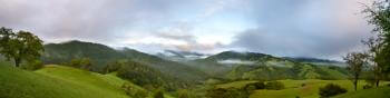 Lush green hills stretching into the distance, covered in green trees, under a light blue morning sky with dark clouds