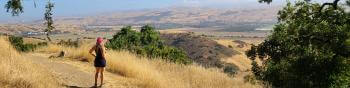 A hiker wearing shorts, tank top, and pink baseball hat stands on a dirt trail looking at a view of a golden valley and open sky