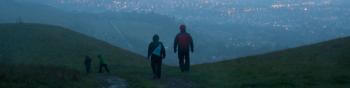A pair of hikers wearing raincoats walks away from the camera down a wet and muddy trail, with city lights below them through a misty haze