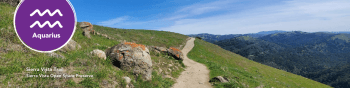 Sierra Vista trail with green hills, a path, and mountains in the back. Aquarius symbol in the left corner.