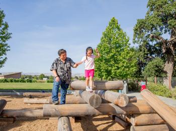 A child wearing pink shorts, a white shirt, and purple ear protection headphones walks across a play structure make up of logs, holding the hand of her father who is wearing jeans and a black and gray shirt. Behind them is a grassy park and blue sky.