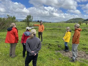 Six people in rain jackets stand in a loose circle in a green meadow, looking towards the left where the person in the center of the image is pointing