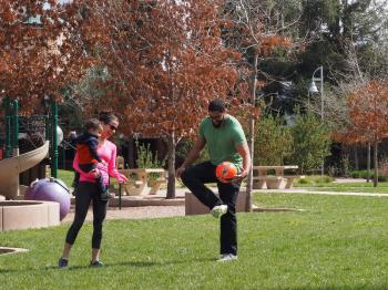 A woman wearing a pink shirt and sunglasses and holding a toddler on her hip smiles and points at the man next to them wearing glasses and a green shirt and juggling an orange soccer ball, at a small park lawn next to a play structure