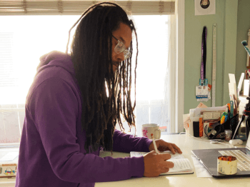 A side profile of Robert Liu-Trujillo shows him working at a desk with a pencil in his hand sketching on a notepad.