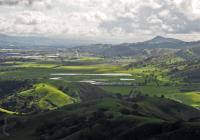 Looking across a lush green landscapes of hills, a large valley, fields, and small bodies of waters, towards mountains in the distance, under a cloud-filled sky