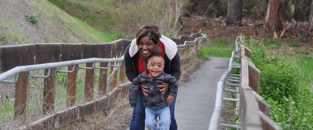 Small boy with mother holding him from behind on a paved park trail both smiling at camera 