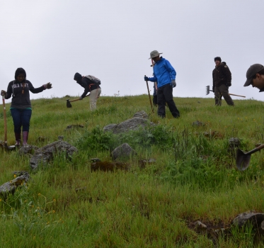 Seven volunteers with jackets and hoes clearing weeds on a green hillside under an overcast sky