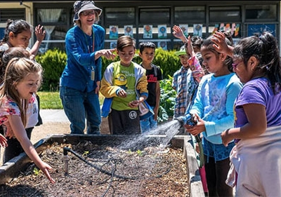 Smiling students and teacher around raised garden bed with sprinkler hose