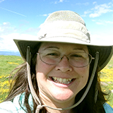 Teri Rogoway wearing glasses and a wide-brimmed hat smiling at the camera, with a field of wildflowers and blue sky behind her