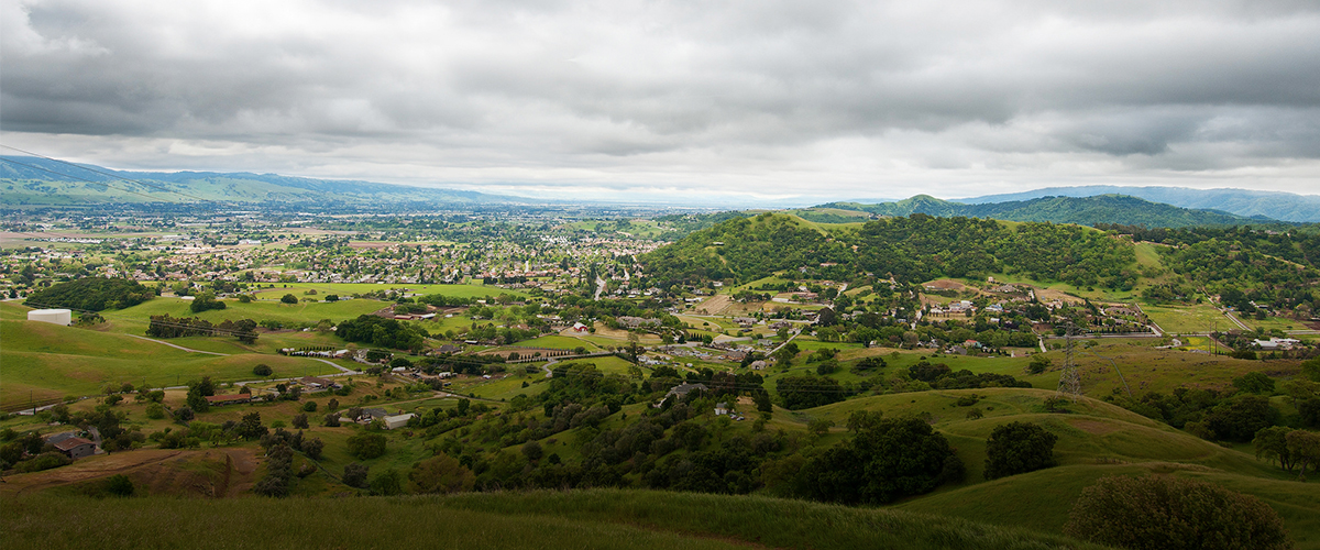 Wide view looking down at houses scattered across the valley and nestled in green hillsides, mountains in distance